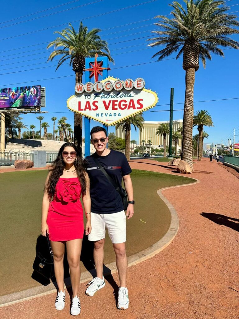 Casal posando ao lado do famoso letreiro de "Welcome to Fabulous Las Vegas", com céu azul e clima desértico ao fundo.